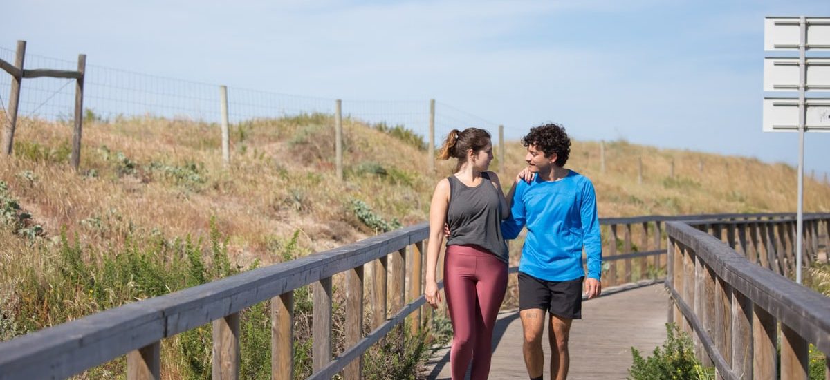 A couple in activewear strolling together on a sunny boardwalk in a natural setting.